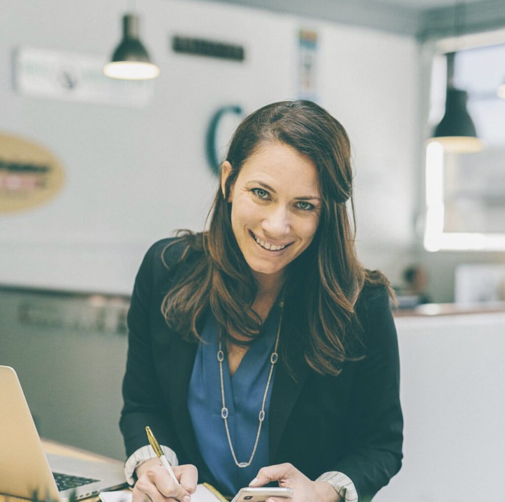 women in blazer and denim shirt looks up smiling from computer ellen grace marketing 